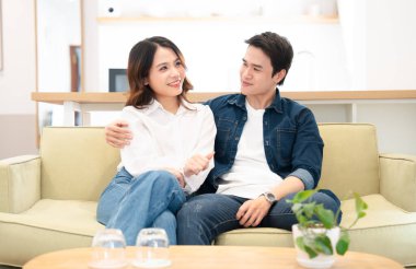 Asyalı bir çift açık yeşil bir kanepede rahatlar ve evde birbirlerine sevgiyle bakarlar.