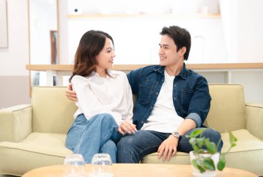 Mutlu genç bir çift, ışık dolu evlerinde bir kanepede kucaklaşır rahat ve mutlu bir şekilde gülümserler.