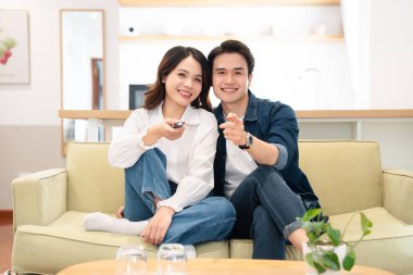 Gülümseyen genç bir çift yeşil bir kanepede oturuyor. Parlak oturma odalarında uzaktan kumandayla.