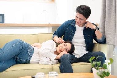 Genç, gülümseyen bir kadın, parlak bir oturma odasındaki kanepede bir erkeğin kucağında rahatça dinlenir.