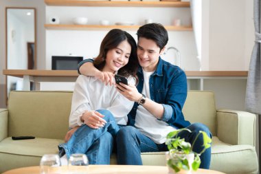 Mutlu bir çift kanepede oturup gülümsüyor ve rahat bir oturma odasında akıllı telefona bakıyor.