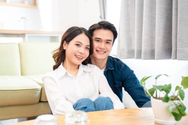 Neşeli bir çift, parlak bir oturma odasında yerde birlikte oturur ve kameraya içten bir şekilde gülümserler.