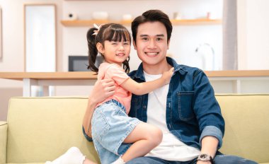 Mutlu bir baba ve küçük kızı kanepede oturmuş kameraya gülümsüyor.