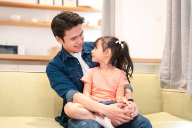 Bir baba ve kızı, aydınlık bir yaşam alanında bir kanepede birlikte oturarak hassas bir anı paylaşıyor.