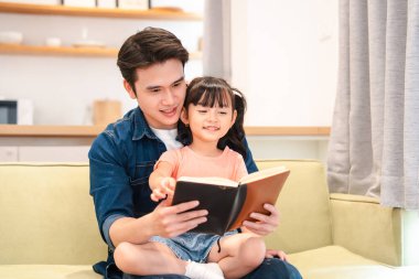 Bir baba ve küçük kızı kanepede oturup kitap okuyarak rahat bir evde güzel vakit geçiriyorlar.