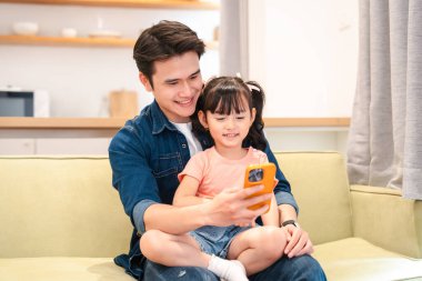 Bir baba ve kızı koltukta oturup cep telefonu ekranına bakarak eğleniyorlar.