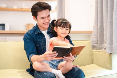 Gülümseyen bir baba, kızına kitap okurken, o da rahat bir odada kucağında oturuyor.