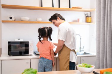 Önlüklü bir baba, kızının modern aletler ve dekorlarla aydınlık bir mutfakta yemek yapmasına yardım eder.