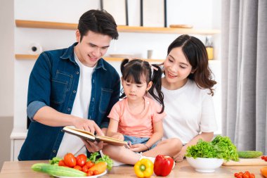 Mutfakta taze sebzelerle çevrili bir yemek tarifi kitabına bakan bir aile salata yapmaya hazır.