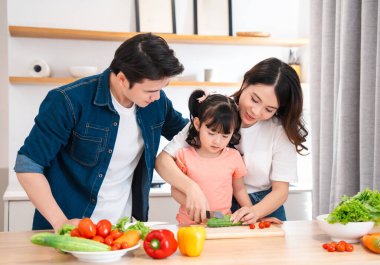 Parlak bir mutfakta aile, küçük bir çocuğa nasıl sebze kesileceğini öğretir.