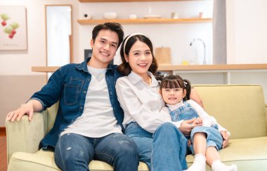 Üç kişilik genç bir aile, rahat bir evde, kanepede yan yana oturmuş gülümsüyor.