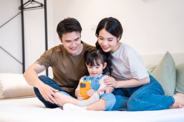 Özenli genç bir aile, kapalı bir yatakta yan yana otururken hücresel bir cihaz kullanmaktan mutluluk duyar.