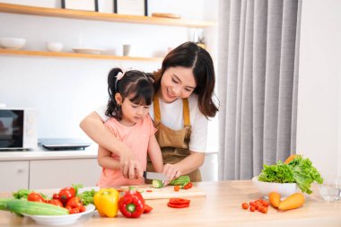 Bir anne genç kızına parlak bir mutfakta tahta kesme tahtasıyla sebze dilimleme konusunda rehberlik eder.