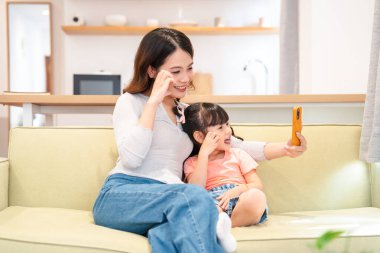 Gülümseyen bir anne ve kız, parlak, modern oturma odalarında bir kanepede selfie çekiyorlar.