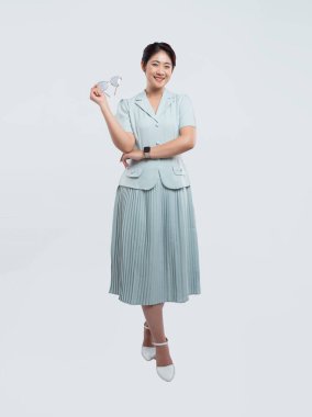 Zarif giyimli bir kadın, açık mavi bir takım elbise ve elinde gözlük tutan beyaz ayakkabılarla güvenle gülümsüyor.