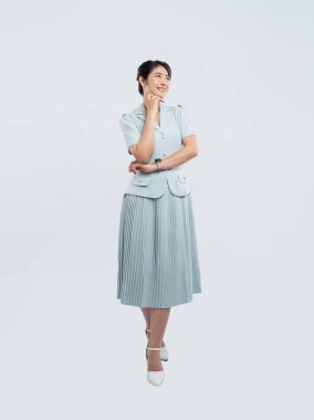 Açık mavi takım elbiseli ve pileli etekli gülümseyen bir kadın düşünceli bir şekilde beyaz bir arkaplana poz veriyor.