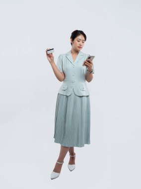 Mavi pileli elbise giymiş genç bir kadın elinde bir kartla telefonuna bakıyor.