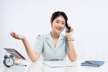 Asyalı kadın masasında ofis malzemeleri ve tabletle telefonuyla konuşurken gülümsüyor.