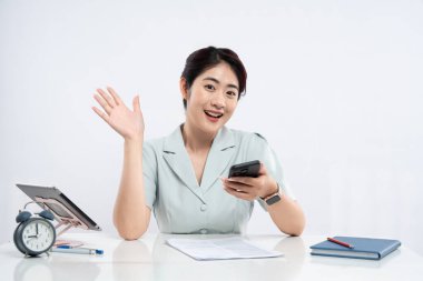 Gülümseyen bir kadın, elinde evrak ve aletlerle beyaz bir masada akıllı telefon tutarken el sallıyor.