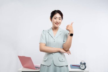 Açık mavi takım elbiseli, gülümseyen bir kadın, parlak, temiz ofis ortamında baş parmağını kaldırıyor.