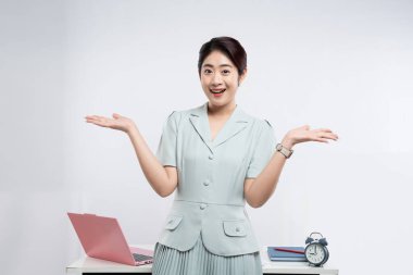 Genç bir kadın gülümsüyor ve avuçları açık bir şekilde elinde dizüstü bilgisayarı ve saati olan bir masada duruyor.