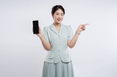 Pastel elbiseli, gülümseyen bir kadın beyaz arka plana karşı bir yandan işaret ederken bir akıllı telefon sunar.