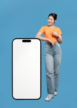 Gündelik kıyafetli neşeli bir kadın mavi arkaplana karşı büyük bir akıllı telefon ekranını işaret ediyor.