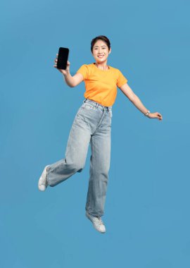 Gülümseyen bir kadın havada zıplarken elinde sağlam mavi bir arka planı olan bir cep telefonu tutuyor.