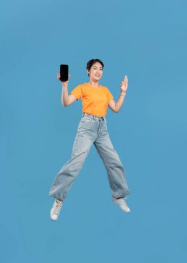 Neşeli genç bir kadın mavi bir arka planın önüne atlıyor ve cep telefonunu gösteriyor.