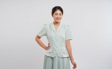 Açık yeşil takım elbiseli ve pileli etekli gülümseyen bir kadın, basit beyaz bir arka plana karşı poz verir.