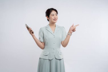 Açık yeşil takım elbiseli gülümseyen Asyalı bir kadın elinde bir telefon tutuyor ve arka planda açık bir yer gösteriyor.