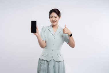 Gülümseyen bir kadın, düz bir arkaplana başparmak atarken akıllı telefon sunar.