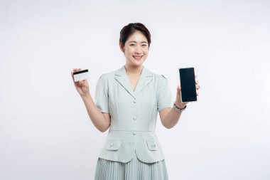 Açık mavi elbiseli, gülümseyen kadın beyaz arka planda elinde kredi kartı ve akıllı telefon tutuyor.