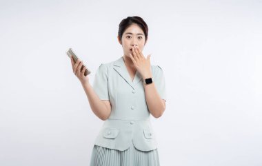 Asyalı bir kadın bir elinde telefon tutarken ve ağzını şokla kapatırken şaşırmış görünür.