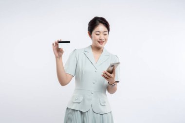 Açık renkli takım elbiseli gülümseyen kadın elinde kredi kartı ve akıllı telefonuyla muhtemelen internetten alışveriş yapıyordur.