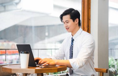 Genç adam iş için giyinmiş bir kahve fincanıyla parlak bir pencerenin önünde dizüstü bilgisayarda çalışıyor.