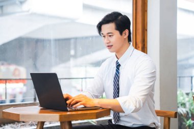 Diz üstü bilgisayarıyla çalışan bir adam, parlak, rahat bir kafe ortamında büyük bir pencerenin yanındaki masada çalışıyor.