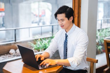 Gömlek ve kravatlı genç profesyonel, dizüstü bilgisayarında çalışıyor. Kafesi doğal ışık ve bitkilerle dolu.