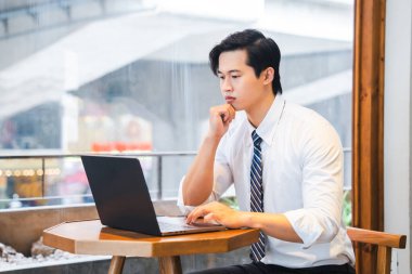 Koyu renk saçlı genç adam pencerenin kenarında dizüstü bilgisayarla oturuyor. Çok odaklanmış görünüyor. Kravat takıyor.