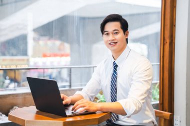 Gülümseyen bir adam dizüstü bilgisayar kullanarak masada oturur. Gömlek ve kravat takar ve yüzünü öne bakar.