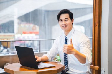 Gömlek ve kravat takan genç bir adam dizüstü bilgisayarla masaya oturur ve baş parmak hareketi yapar.