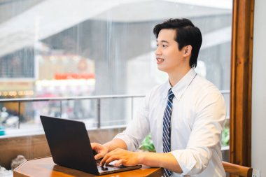 Genç profesyonel bir adam bir kafede dizüstü bilgisayarıyla çalışır pencereden bakar ve düşünceli bir şekilde gülümser.