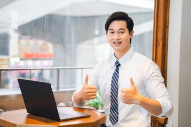 Harika gülümseyen bir adam dizüstü bilgisayarla otururken iki parmağını kaldırır. Çok başarılı görünüyor.