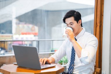 Profesyonel bir adam, modern bir çalışma alanının penceresinin yanındaki kağıt bardaktan içerken dizüstü bilgisayarda çalışır.