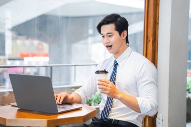 Genç profesyonel elinde kahve tutarken dizüstü bilgisayarda çalışıyor. Pencerenin yanında oturuyor.