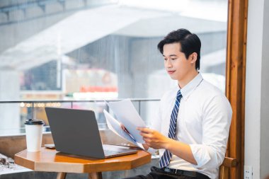 Profesyonel bir adam bir kafede iş kıyafeti giymiş rahat bir atmosferde dizüstü bilgisayar kullanarak evrakları inceliyor.