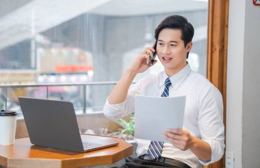 Kafe ortamında Asyalı bir işadamı elinde laptopuyla telefonuyla konuşuyor.