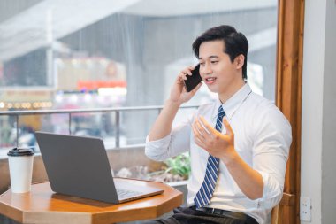 Kafede dizüstü bilgisayarla çalışan profesyonel bir adam cep telefonuyla gömlek ve kravat takarak konuşuyor.