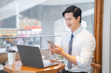 Yakalı gömlekli ve kravatlı genç işadamı cep telefonunu ve bilgisayarını parlak bir kahve dükkanında kullanıyor.