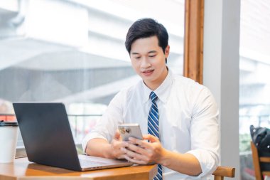 Bir adam önünde dizüstü bilgisayar ve kahve fincanı olan bir masada otururken telefon kullanıyor.